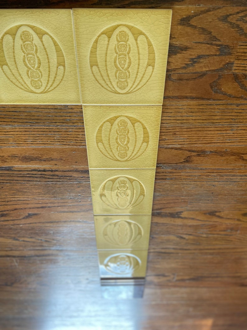 May include: A row of six yellow ceramic tiles with a repeating pattern of a stylized flower design. The tiles are arranged vertically and are set against a wood background.