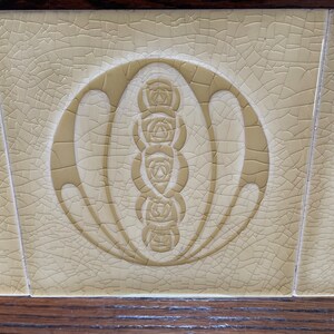 May include: A close-up of a cream-colored ceramic tile with a crackled glaze. The tile features a stylized design of a circle with a series of five stylized roses inside.