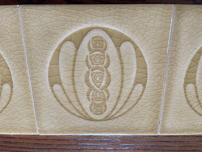 May include: A close-up of a cream-colored ceramic tile with a crackled glaze. The tile features a stylized design of a circle with a series of five stylized roses inside.