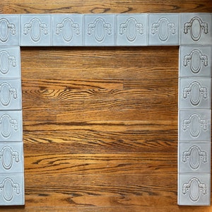 May include: A set of gray ceramic tiles with a decorative design of a wreath and oval. The tiles are arranged in a rectangular shape, forming a fireplace surround.