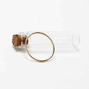 Peut inclure: Une bague en or avec un design rond simple. La bague est pos&eacute;e sur une surface blanche &agrave; c&ocirc;t&eacute; d'un flacon en verre transparent avec un bouchon en li&egrave;ge.