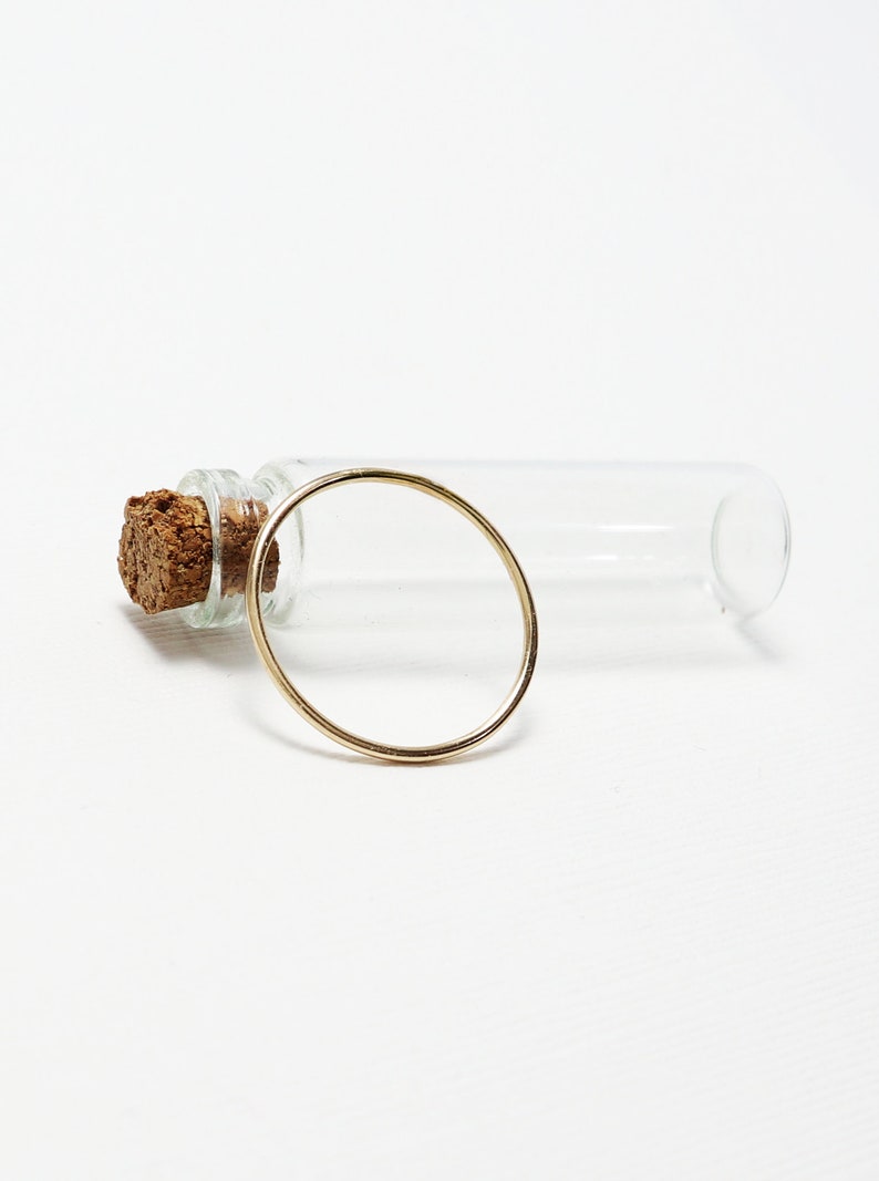 Peut inclure: Une bague en or avec un design rond simple. La bague est pos&eacute;e sur une surface blanche &agrave; c&ocirc;t&eacute; d'un flacon en verre transparent avec un bouchon en li&egrave;ge.