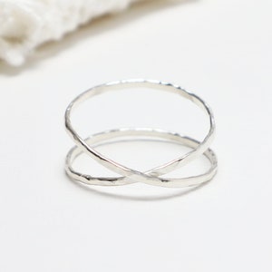 May include: A silver ring with a unique crisscross design. The ring features two bands that intersect, creating an X shape. The metal has a textured, hammered appearance, adding visual interest. The ring is displayed on a white surface.