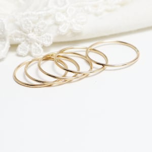 May include: Five gold stacking rings, each with a simple, smooth, circular design. The rings are arranged in a stack, with the largest ring at the bottom and the smallest ring at the top.