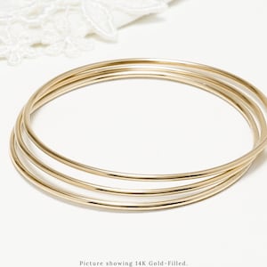 May include: Three gold bangles stacked on top of each other. The bangles are available in 14K gold-filled, 14K rose gold-filled, and sterling silver.