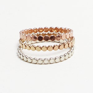 May include: Three delicate stacking rings made of silver, gold, and rose gold. Each ring is composed of small, round beads.