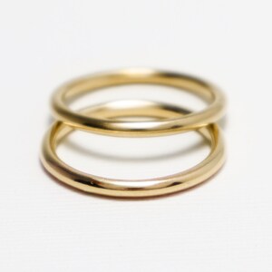 May include: Two gold rings, one atop the other, against a white backdrop. The rings have a smooth, rounded design and a shiny finish, reflecting light. The rings appear to be made of metal.