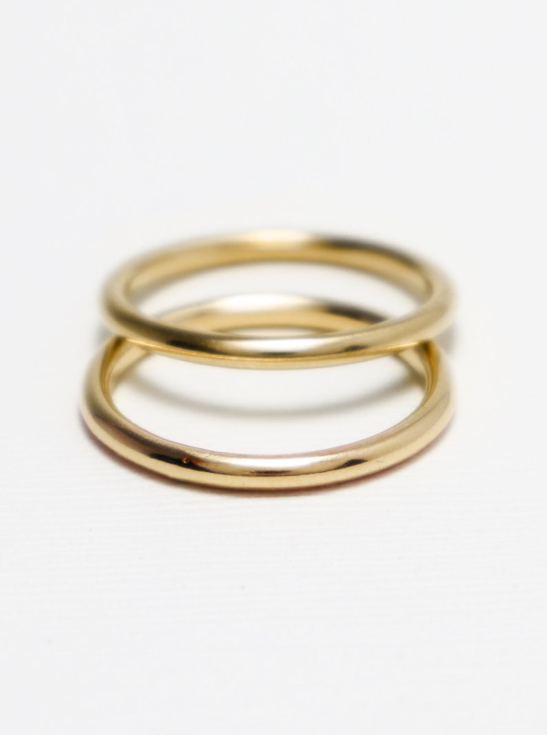 May include: Two gold rings, one atop the other, against a white backdrop. The rings have a smooth, rounded design and a shiny finish, reflecting light. The rings appear to be made of metal.