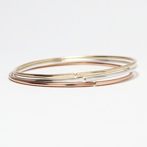 May include: Three thin metal bangles stacked on top of each other. The top bangle is gold, the middle bangle is silver, and the bottom bangle is rose gold.