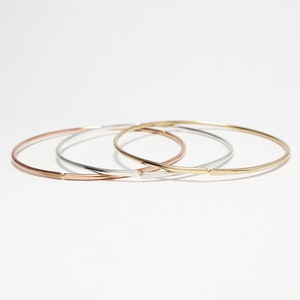 May include: Three thin bangle bracelets in gold, silver, and rose gold. The bracelets are stacked on top of each other.