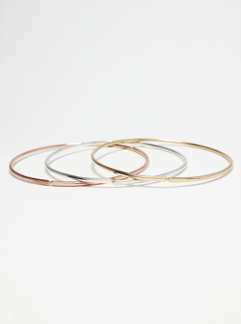 May include: Three thin bangle bracelets in gold, silver, and rose gold. The bracelets are stacked on top of each other.