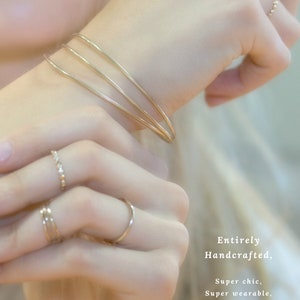 May include: Three gold bangle bracelets stacked on a wrist. The bracelets are simple and delicate. The image also shows three gold rings on a finger. The rings are also simple and delicate. Entirely Handcrafted, Super chic, Super wearable, Made just for you! Rings & Bracelets available at IrresistiblyMinimal.etsy.com