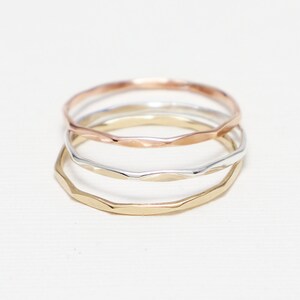 May include: Three thin gold rings stacked on top of each other. The top ring is rose gold, the middle ring is silver, and the bottom ring is yellow gold. All three rings have a hammered texture.