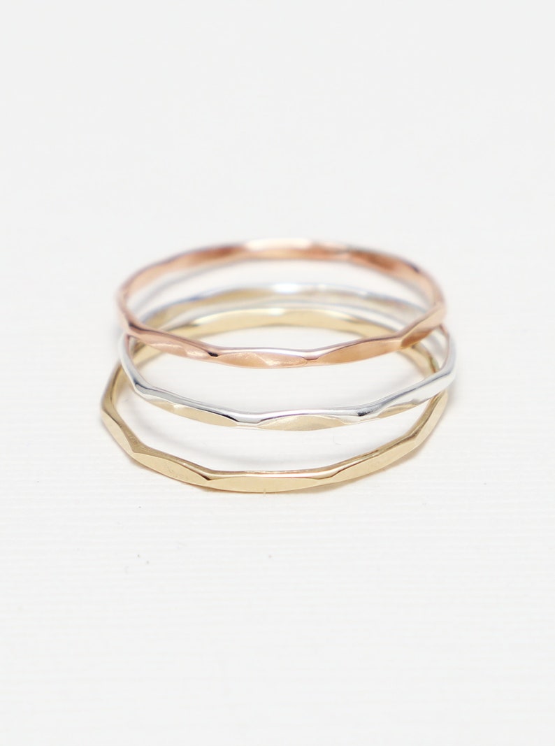 May include: Three thin gold rings stacked on top of each other. The top ring is rose gold, the middle ring is silver, and the bottom ring is yellow gold. All three rings have a hammered texture.