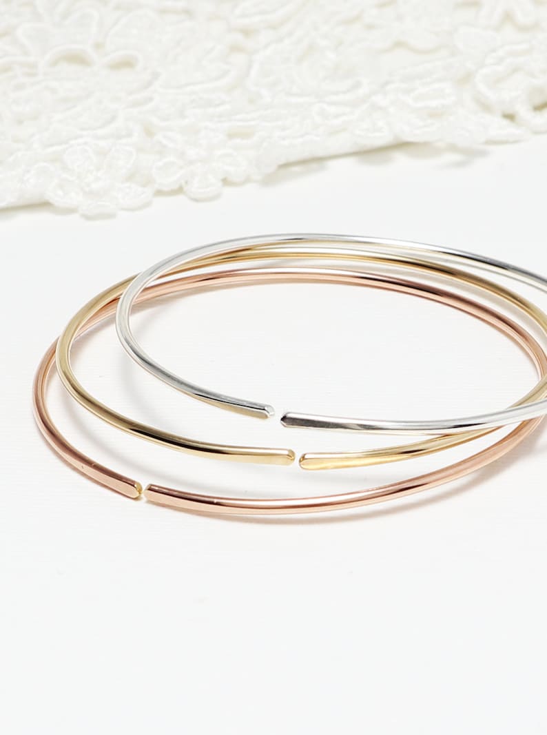 May include: Three simple bangle bracelets in different metal finishes: rose gold, gold, and silver. The bracelets are stacked on top of each other.