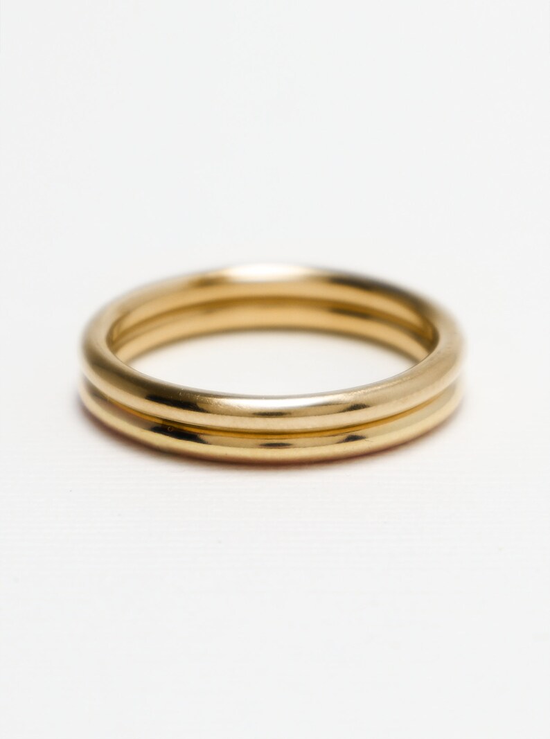 May include: Two gold-coloured rings, possibly metal, are shown against a white backdrop. The rings have a smooth, shiny surface and a simple, classic design. These are likely jewellery items.