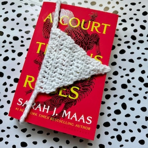 May include: A white crocheted triangle bookmark with a long tail is placed on a red book titled "A Court of Thorns and Roses" by Sarah J. Maas. The book cover features a black and red illustration of a mythical creature.