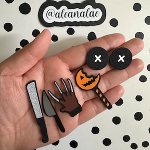 May include: A set of Halloween-themed felt patches, including a jack-o'-lantern lollipop, a knife, a Freddy Krueger-style glove, and two black circles with an 'X' on each. The patches are on a hand against a black and white polka dot background.