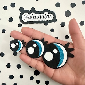 Puede incluir: Tres ojos saltones de fieltro negro con pupilas blancas y detalles de purpurina azul. Los ojos son de diferentes tamaños y se sostienen en una mano.