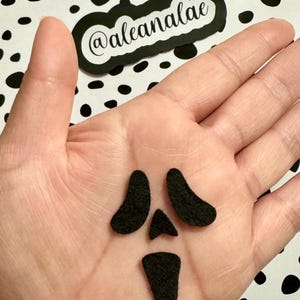 May include: A black felt ghost face cutout with two eyes, a nose, and a mouth. The ghost face is on a person's hand.