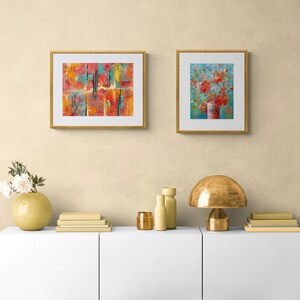 May include: Two framed abstract paintings on a white wall above a white shelf with a gold lamp, two gold vases, and a stack of books. The painting on the left is a colorful abstract design with red, orange, yellow, and blue colors. The painting on the right is a blue background with a red flower bouquet in a vase.