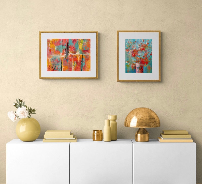 May include: Two framed abstract paintings on a white wall above a white shelf with a gold lamp, two gold vases, and a stack of books. The painting on the left is a colorful abstract design with red, orange, yellow, and blue colors. The painting on the right is a blue background with a red flower bouquet in a vase.