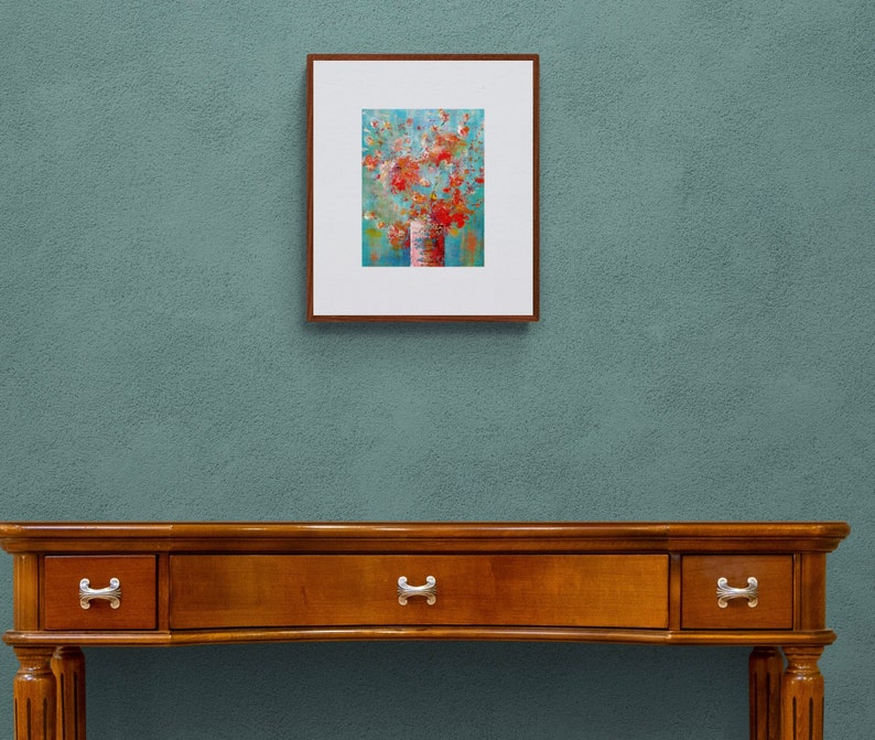 May include: A framed painting of a bouquet of red flowers in a vase against a blue background. The painting is hanging on a wall above a wooden table with drawers.