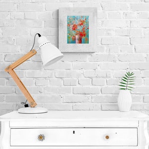 May include: A white lamp with a wooden arm sits on a white dresser with a drawer. A framed painting of a floral arrangement in a vase is on the wall behind the lamp. A small potted plant is on the dresser.