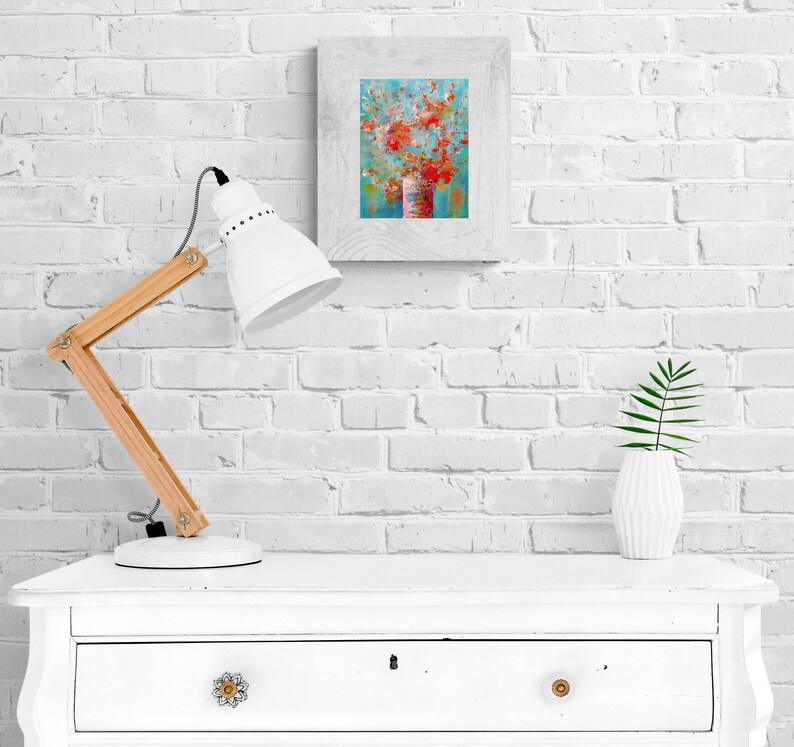 May include: A white lamp with a wooden arm sits on a white dresser with a drawer. A framed painting of a floral arrangement in a vase is on the wall behind the lamp. A small potted plant is on the dresser.