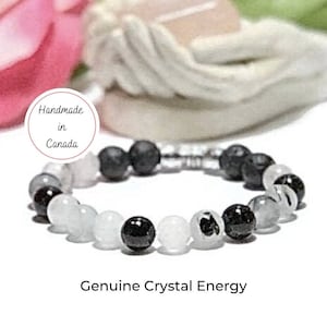 May include: A black and white crystal bracelet with a silver metal connector. The bracelet features black lava beads, white agate beads, and black onyx beads. The bracelet is handmade in Canada and features the text "Genuine Crystal Energy".