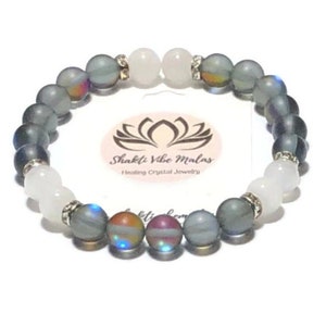 May include: A gray and white beaded bracelet with iridescent accents. The bracelet is made with semi-precious stones and features a silver charm with the text "Shakti Vibe Malas Healing Crystal Jewelry".