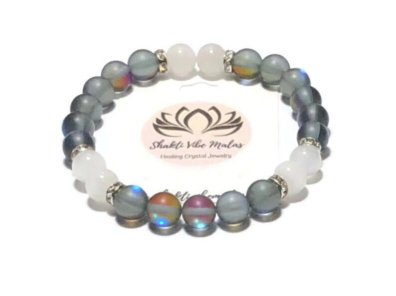 May include: A gray and white beaded bracelet with iridescent accents. The bracelet is made with semi-precious stones and features a silver charm with the text "Shakti Vibe Malas Healing Crystal Jewelry".