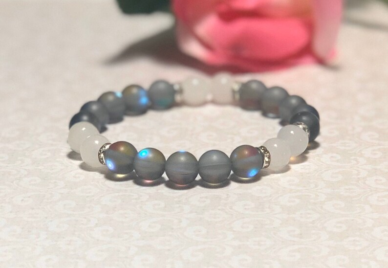 May include: A gray and white beaded bracelet with iridescent beads. The bracelet is made with natural stones and has a silver clasp.