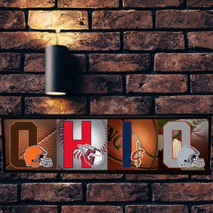 Puede incluir: Un letrero de madera con la palabra "OHIO" escrita en letras grandes. Cada letra está decorada con el logotipo de un equipo deportivo diferente: un casco de fútbol americano, una pelota de béisbol, un balón de baloncesto y un palo de hockey. El letrero es una excelente manera de mostrar tu espíritu de equipo.