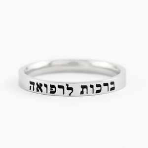 May include: A polished silver-toned band ring featuring a smooth, flat outer surface. The ring is engraved with black Hebrew text that reads "ברכות לרפואה" which translates to blessings for healing, centered along the front of the metal band.