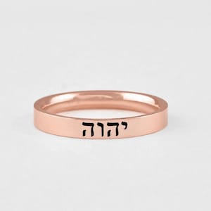 May include: A rose gold band ring featuring a flat outer surface engraved with the Hebrew characters יהוה in black ink. The ring has a smooth, polished finish and a consistent width throughout its circular design, presenting a clean and simple aesthetic.