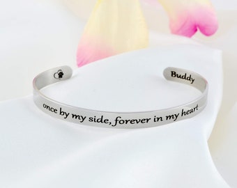May include: A silver-toned metal cuff bracelet featuring the engraved text "once by my side, forever in my heart" on the outer band. The inner band displays the name "Buddy" and a small heart icon with a paw print inside.