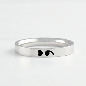 May include: A polished silver-toned band ring featuring a small, black, engraved heart and semicolon symbol on the outer surface. The ring has a smooth, flat profile and a reflective metallic finish, showcasing a clean and simple design.