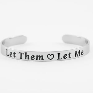May include: A silver-colored cuff bracelet with the words "Let Them ♥ Let Me" engraved in black. The bracelet has an open design and is displayed against a white background.
