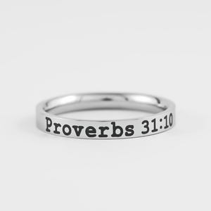 May include: A silver-colored ring with the words "Proverbs 31:10" engraved in black. The ring is thin and has a smooth, polished surface. The text is centered on the ring.