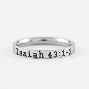 May include: A silver-colored ring with the inscription "Isaiah 43:1-2" in black lettering. The ring is a simple band design, likely made of stainless steel or a similar metal. The text is centered on the ring's exterior.