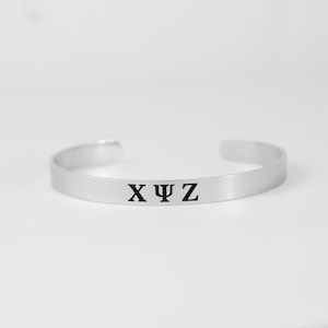 May include: A silver-toned metal cuff bracelet featuring a smooth, polished finish. The center of the band is engraved with the black Greek letters Chi, Psi, and Zeta in a clear, bold serif font. The bracelet has an open-ended design.