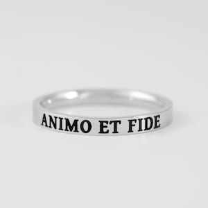 Peut inclure: Une bague anneau en métal argenté poli avec le texte noir gravé ANIMO ET FIDE centré sur la surface extérieure. La bague présente un profil plat et lisse, avec une largeur constante sur toute la circonférence de l'anneau métallique.