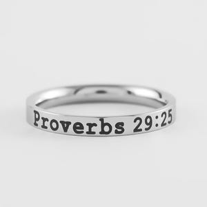 May include: A silver-colored ring with the inscription "Proverbs 29:25" in black lettering. The ring is a simple band design, likely made of stainless steel or a similar metal. The text is centered on the ring's exterior.
