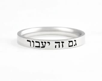 May include: A silver-toned metal ring with a smooth, polished finish. The outer surface of the band features black engraved Hebrew text that reads "גם זה יעבור," which translates to "this too shall pass," in a clear, sans-serif typeface.