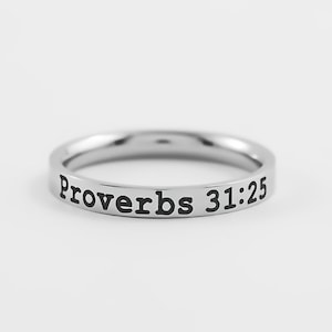 May include: A silver-colored ring with the inscription "Proverbs 31:25" in black lettering. The ring is circular and appears to be made of metal, possibly stainless steel. The text is centered around the band.