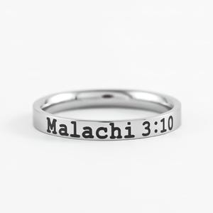 May include: A silver ring with the text "Malachi 3:10" engraved in black. The ring is circular and sits on a white background. The ring is a simple band.