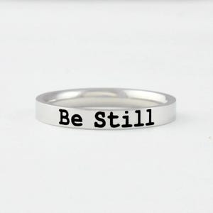 May include: A polished silver-toned metal band ring featuring a smooth, flat surface. The words "Be Still" are engraved in a clear, black, sans-serif font along the outer circumference of the ring, creating a simple and clean aesthetic design.