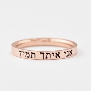 May include: A rose gold band ring featuring a flat outer surface engraved with black Hebrew text that reads "אני איתך תמיד". The ring has a smooth, polished metallic finish and a consistent, slim width throughout the entire circular band structure.