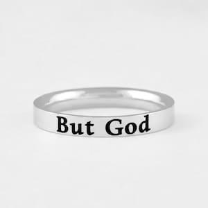 Peut inclure: Une bague anneau en métal argenté poli avec le texte But God gravé en police serif noire sur la surface extérieure. La bague présente un profil plat et lisse avec une largeur constante, offrant un design simple et épuré.
